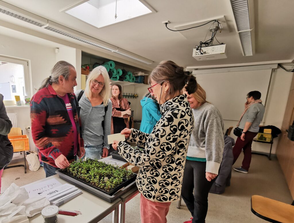 Zwei Menschen stehen sich an einem Tisch gegenüber auf dem sich Saatgut befindet. Die Menschen unterhalten sich. Im Hintergrund sind weitere Menschen zu erkennen, die umherlaufen.